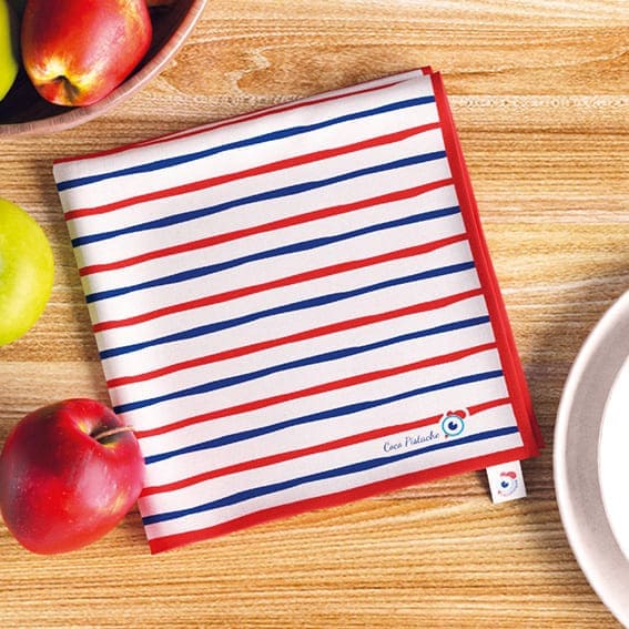 Serviette en tissu La Marinière de la famille Coco Pistache. Ambiance pommes, table en bois, assiettes. Motif marinière coq, signature Coco Pistache. Tricolore, bleu, blanc, rouge. France.