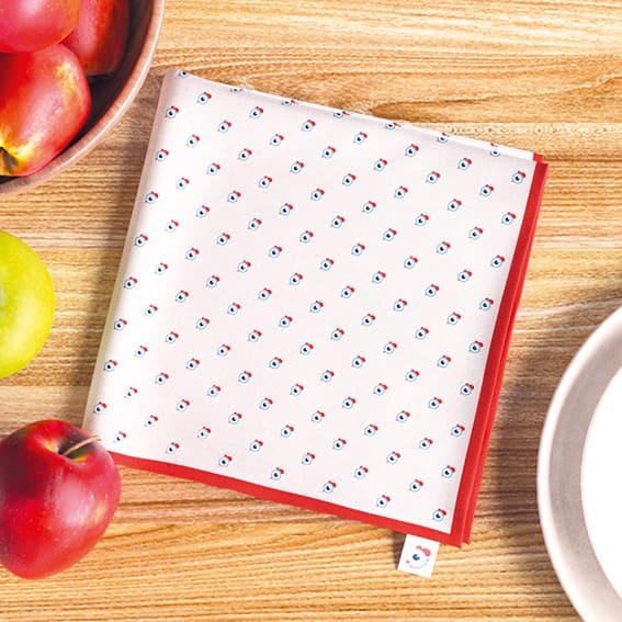 Serviette française La Coquette de la famille Coco Pistache. Ambiance pommes, table en bois, assiettes. Motif pois coq, signature Coco Pistache. Tricolore, bleu, blanc, rouge. France.